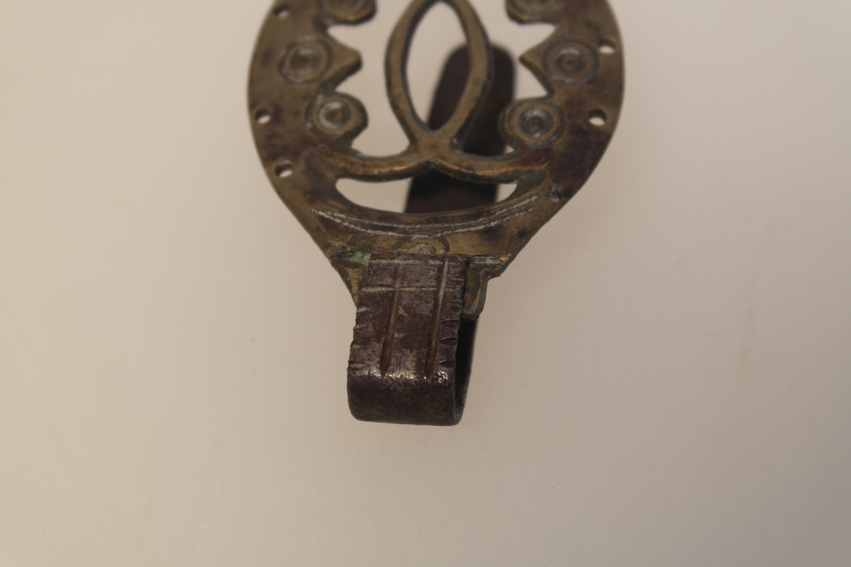 Lommekrok i støpt messing og jern. Gjennombrutt ornament i messingplaten. Kroken har huller, slik at den kan sys fast til lommen.