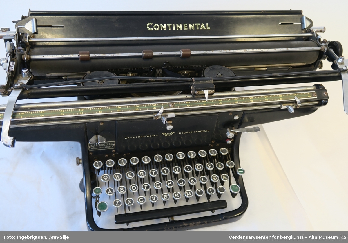 Rektangulær svartlakkert mekanisk skrivemaskin på fire bein med en bred skrivevalse som går langt utover maskinens grunnform. Maskinen har et overtrekk i tekstil.