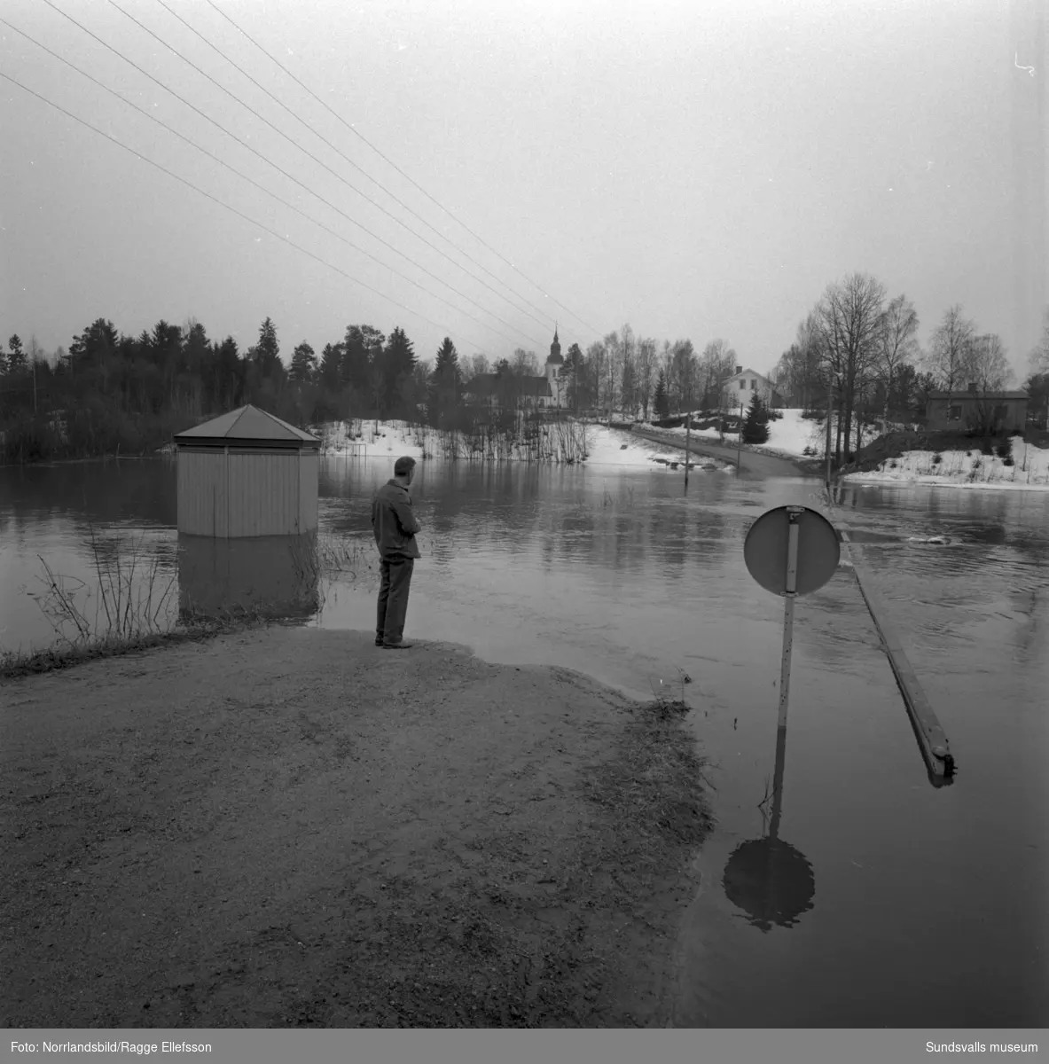 Sättnaån har svämmat över vid Ånäsparken efter den snörika vintern 1966. En man betraktar förödelsen och i bakgrunden syns Sättna kyrka