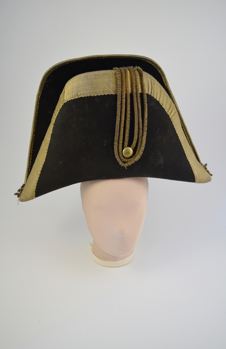 Svart "napoleonshatt"/snutehatt med bret gullbånd langs kanten på for- og bakside. Gullfarget knapp og gullbånd som dekorasjon på hattens forside.
