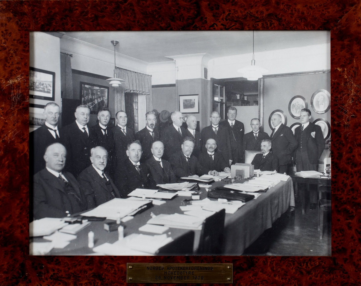Gruppefoto av 19 voksne menn i dress. Sju sittende foran og 12 stående bak. Møtebord i forgrunnen.

Gruppen er Norges Apotekerforenings Hovedstyre i møte 25. november 1929.
