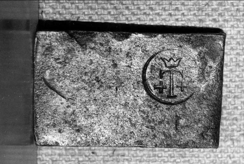 Avhugget stycke av järnband, byggnadsjärn med järnstämpel. Bokstaven T, krona över samt ett plustecken på vardera sidan.