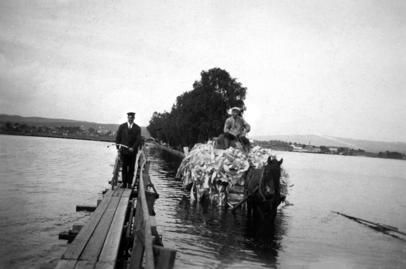 Svart-hvitt foto fra 1927. Gangbru med mann og hestetransport med stort lass som kjører på ordinær bru i flomvann.