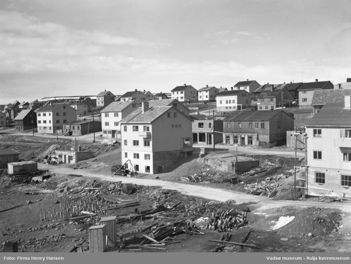 Vadsø 3.6.1951, sentrum. Bildet er tatt fra sørøst mot nordvest, trolig fra et hustak sør for torget. Bildet viser bygninger nord for Kaigata og langs Havnegata, både ferdige og uferdige bygg og brakker. I forkant en del av fjæra sør for Kaigata.