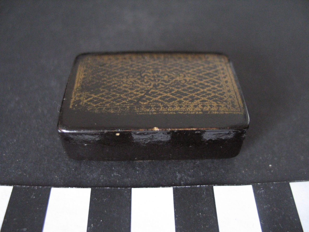 Rektangulär snusdosa av svartlackad papier-maché.
Lock med gångjärn. 
På locket tryckt dekor i form av guldfärgat mönster.