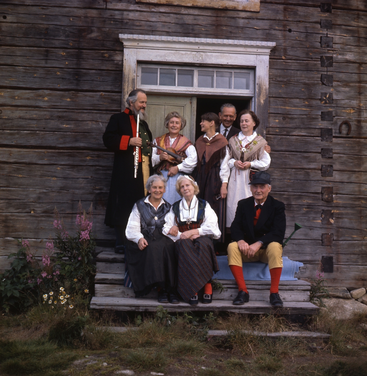 Albert och Lisa Viksten(främre raden från höger), Adéle Mickelsson (bakre raden till höger) med flera ute på en trappa till en timmerbyggnad. De är klädda i hembygdsdräkter. Några håller i musikinstrument.