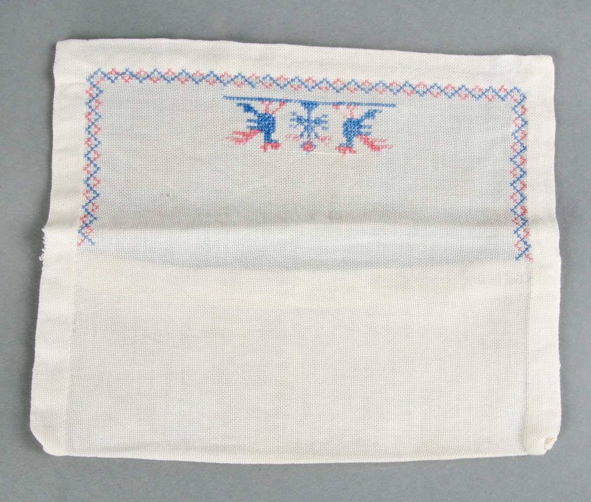 Servettväska av vitt glest tuskaftat bomullstyg handsydd med korsstygnsbroderi i merceriserad bomullstråd, två fåglar och bård i blått och rött.

