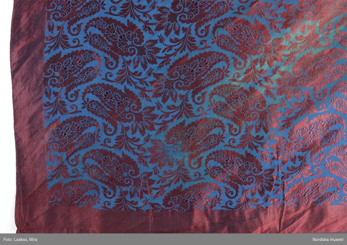 Kvadratiskt halskläde i halvsidensatin med jacquardvävt mönster i vinrött silke på blått bomullsbotten, indiska miribotafigurer över hela ytan med slät vinröd kant runtom. 2 handsydda fållar, 2 stadkanter. 
Tydliga veck efter att ha varit vikt i tresnibb. Har troligen varit en manshalsduk, eftersom den är rätt liten och saknar fransar.
/Berit Eldvik 2011-11-10