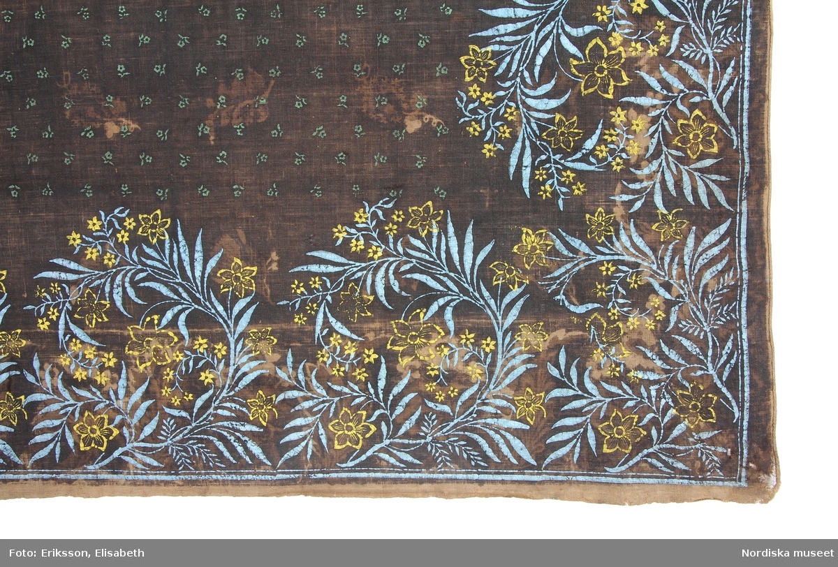 Av  brunsvart bomullslärft, från början ett kattunhalskläde med tryckt blom-och bladmönster i bård och spridda blommor i spegeln, överfärgat svart och på baksidan tryckt med nytt mönster av enklare slag med linoljefärg och tryckstockar. I mittspegeln små strödda gröna blommor, bred kantbård som längs 2 mötande sidor har ett yvigt bladmönster i blått med gula blommor och längs de andra sidorna  har ymnighetshorn med uppväxande prickiga blommor i grönt och vitt och i ytterkant en enkel liten blomranka. Tryckstocken till detta mönster har varit försedd med metallstift som ger det småprickiga utseendet. Båda mönstren var moderna på 1820-30-talet i t.ex. broderimönster men kan ha brukats längre som tryckstockar.  I ena hörnet en märkning med stora stygn som är färgarens märkning av färggodset. 
 Anm. Ursprung okänt, kommer från en samlare som har märkt klädet med numret 2162 i ena hörnet. Möjligen från södra Norrland, Hälsingland, Gästrikland, Dalarna.
Mycket intressant att se hur ett gammalt kattunhalskläde har  återanvänts.
/Berit Eldvik 2012-01