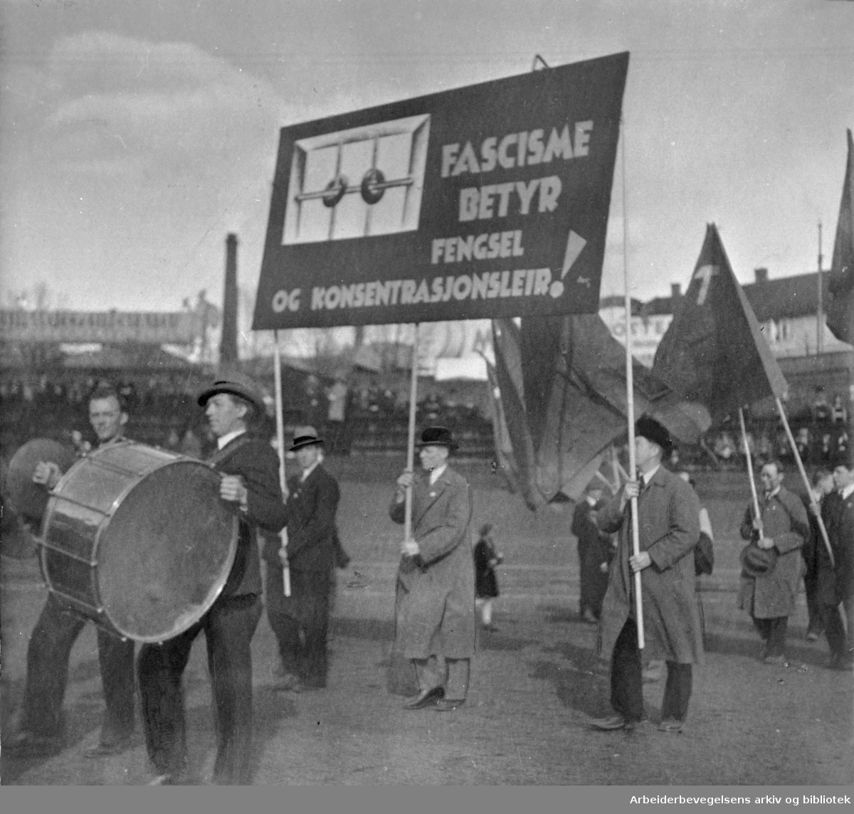 1. mai i Oslo, 1935. Demonstrasjonstoget marsjerer inn på Dælenenga stadion, det avslutningsarrangementet skal holdes. Parole: Fascisme betyr fengsel og konsentrasjonsleir!