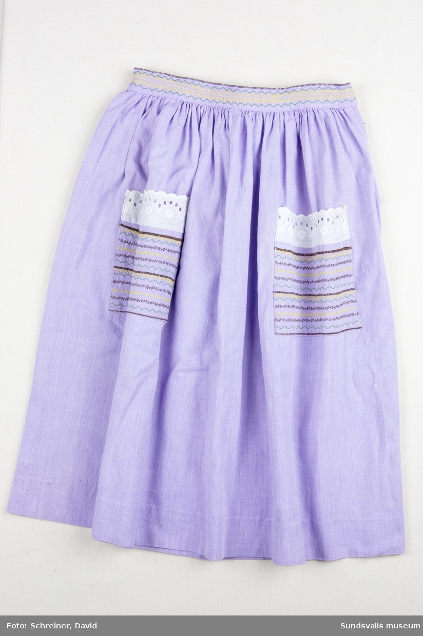 En lila barnkjol med två maskinbroderade och spetskantade fickor. Linningen i midjan bär samma utsmyckning som fickorna. Kjolen har en infälld dragkedja i sidan.