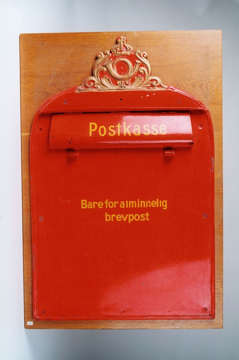 Postkasseskilt/innkast i metall som er rødmalt. Postinnkastet er montert på treplate og har gullfarget posthornemblem og gul skrift.