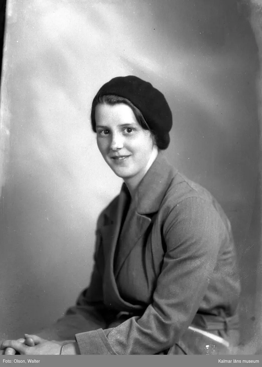 Ateljébild på en kvinna i armbandsur, kappa och mössa. Enligt Walter Olsons journal är bilden beställd av Anna Andersson ifrån gamla lasarettet.