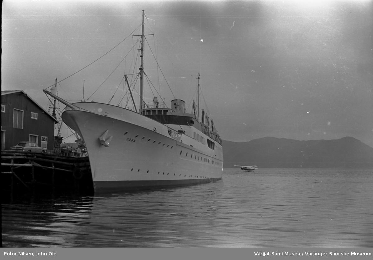 En båt ved navn "Gann" ligger ved kai i Bukta i Alta, et lite sjøfly i bakgrunnen. Alta, Juli 1967