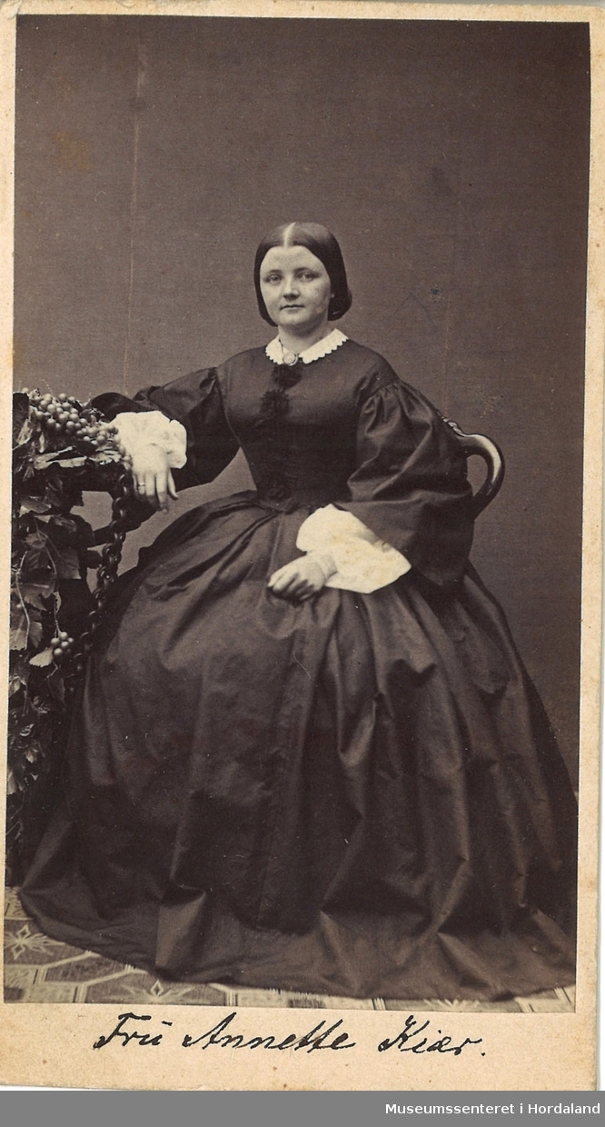 heilfigur fotografi av ung kvinne med stor, vid, langerma kjole med kvit krage og mansjettar som sit på ein stol