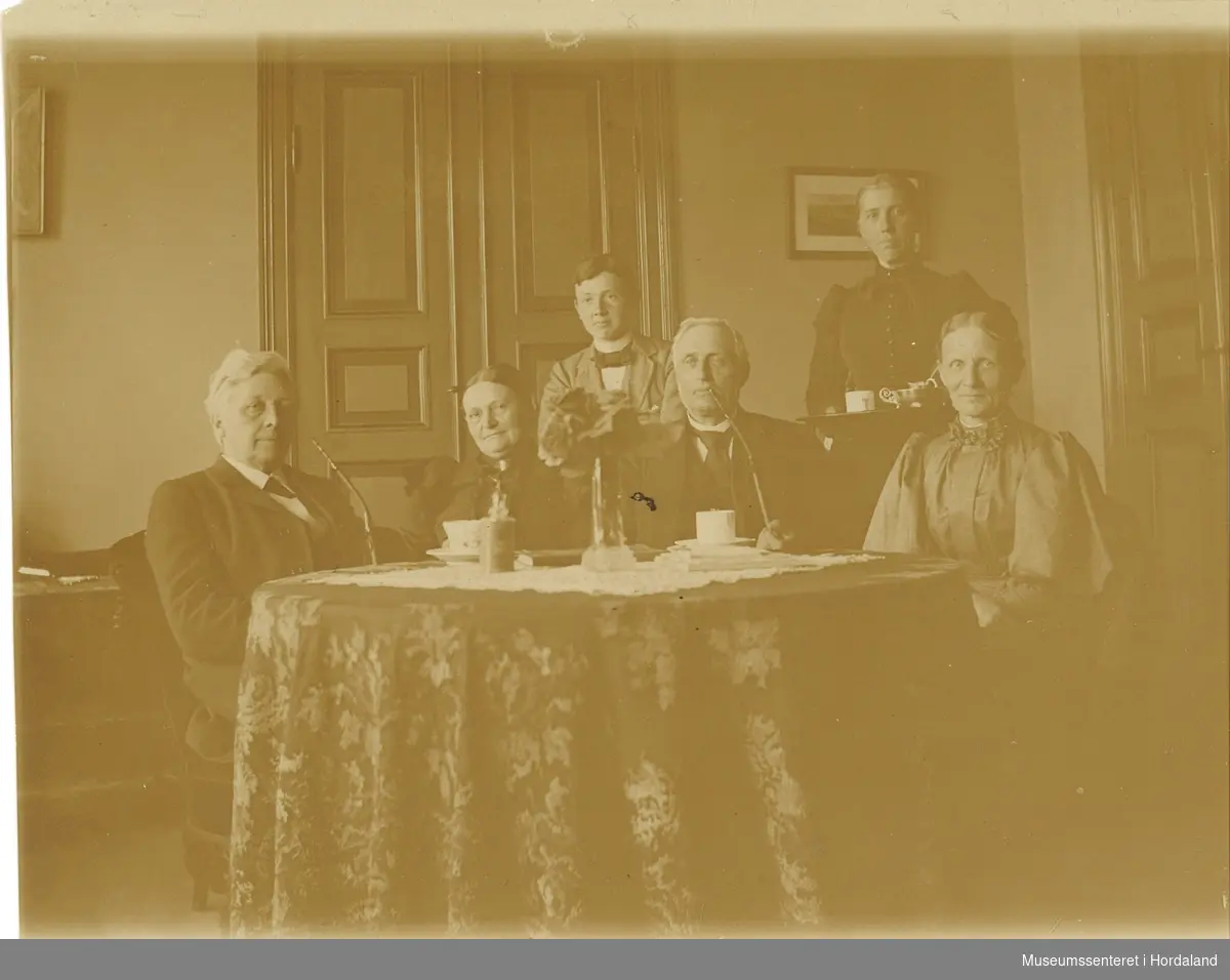 amatørfotografi av to menn, tre kvinner og ein gut som sit og står med eit rundt bord. Frå venstre ser ein Harald og Margrete Arnesen, paret til høgre er truleg Anders og Annette Kiær.