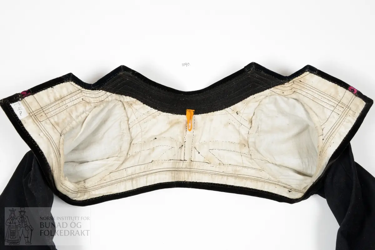 Opentrøye sauma av svart blank glatt klede, lerretsvove. Heilklipt rygg, bandprydd. Oppståande låg krage. Tosaumserme med bryningar. Open ermsliss med 3 stk knappar og knapphol til lukking. Bandpryding av blomstermønstra fløyelsband og svart bomullsfløyel til pryding av krage, framkantar, nedkantar og ermeslissar.  For av kvit litt glissen stiva bomullslerret. Maskinsaum og handsaum.