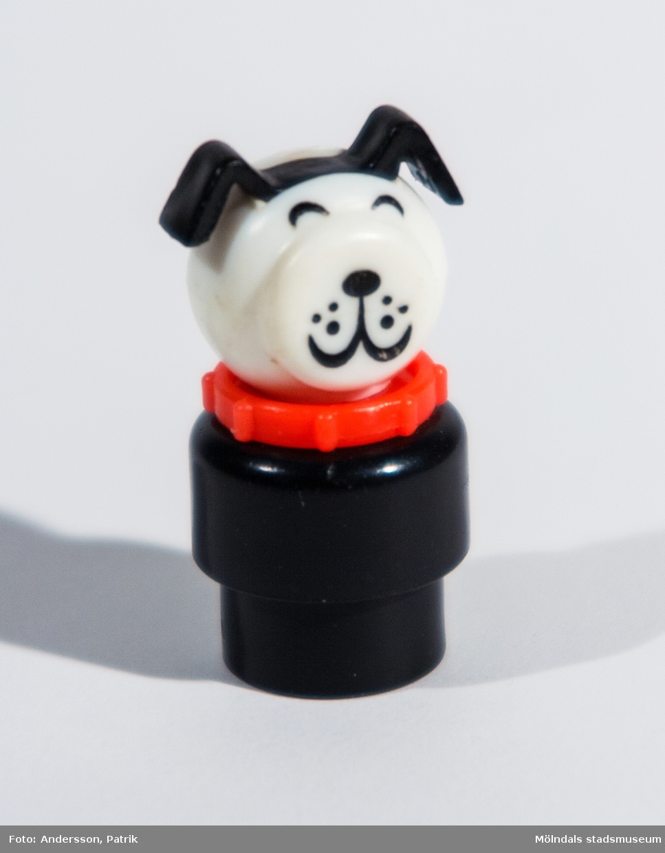 Rund hundfigur av plast tillhörande Fisher-Pricegarage från 1970-talet. "Kroppen" består av två delar varav den nedersta är smalare för att passa ihop med tillhörande leksaksbil. Ihålig underifrån och längst in är "huvudet" fastskruvat. Mellan kropp och huvud sitter ett hundhalsband. "Huvudet" består av en något tillplattad plastkula med markerade hundöron samt målade ögon, morrhår, nos och mun. Inköpt 2001 på Second Hand av givaren, vars barn har lekt med det.