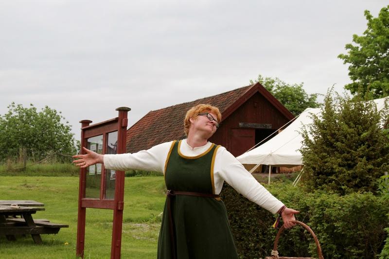 Rødhåret kvinne i middelalderdrakt, med kurv i hånda, slår ut med armene for å ønske velkommen til Urtehagen
