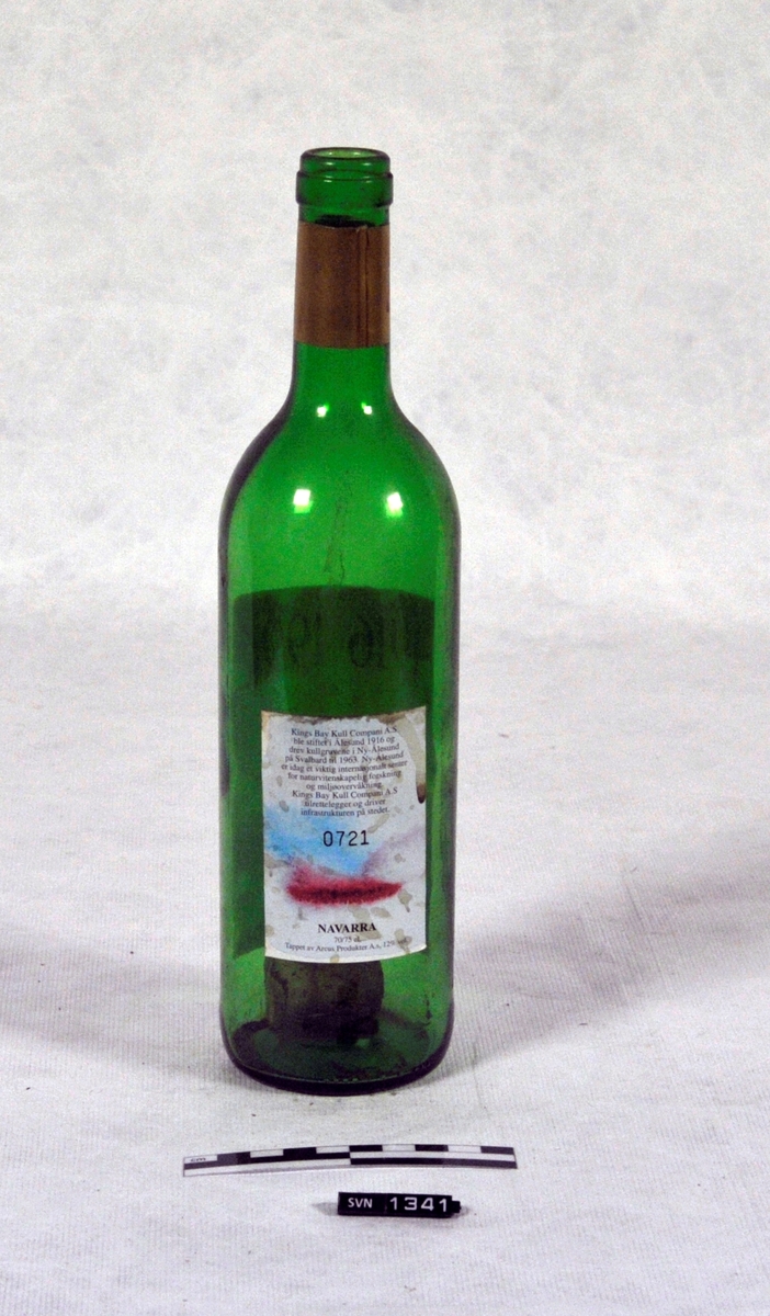 Trekasse med treflis og en fjær.
Kassen inneholder to flasker ( B og C)
Flaskene er jubileumsflasker, en fra Kings Bay Kull Company og en fra Store Norske Spitsbergen Kullkompani.
Kassen er delvis åpen på en side.
Det er en skillevegg mellom de to flaskene.
Flaskene er tomme
Flasken B er av grønt glass med rød/rosa etikett. Flasken er en jubileumsflaske for Store Norske Spitsbergen Kullkompani Flasken har skrukork

Flasken C er av grønt glass med hvit etikett med malt bildetrykk. Flasken er jubileumsflaske for Kings Bay Kull Company. Flasken har ikke kork.