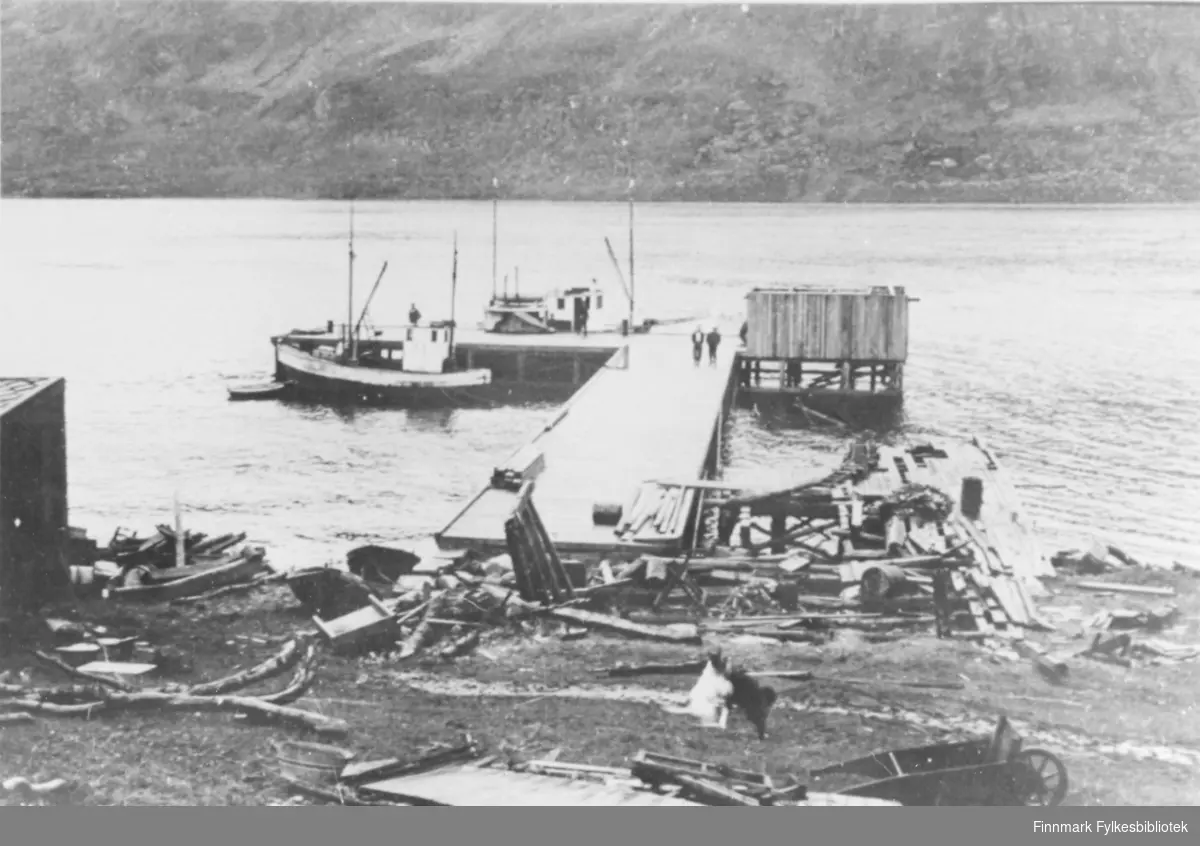 Oversiktsbilde over kai i Maursund. To fiskebåter ligger til kai. I forgrunn av bildet, på land, ligger mye materialer og skrot som er blitt liggende etter krigen. Ytterst på kaia er det reist et skur.