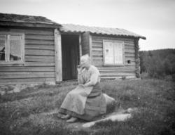 Eldre dame med pipe sitter på en stein ved tømmerstue.