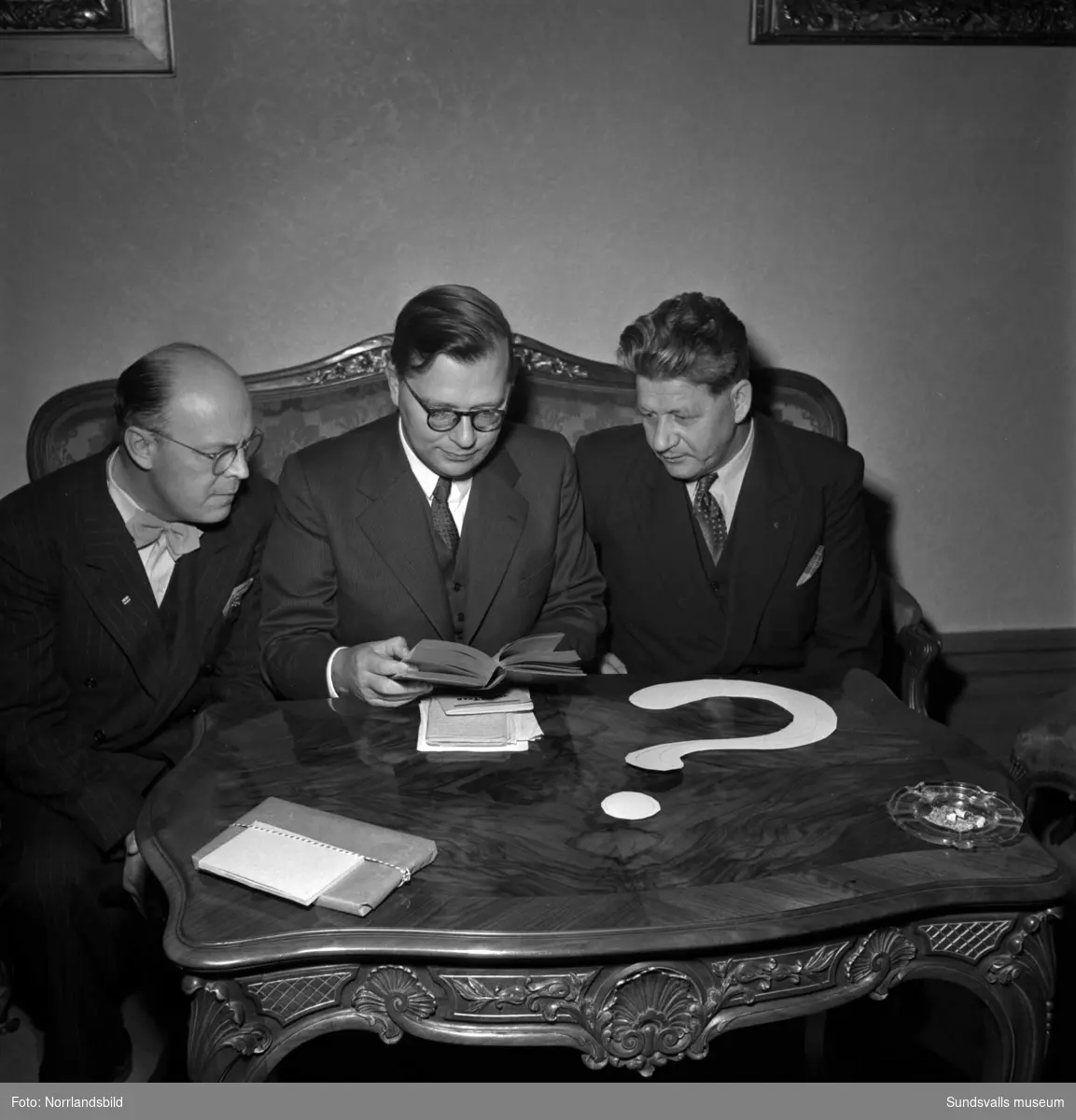 Frågesport i Stadshussalongen, tre män runt ett bord med ett frågetecken på.