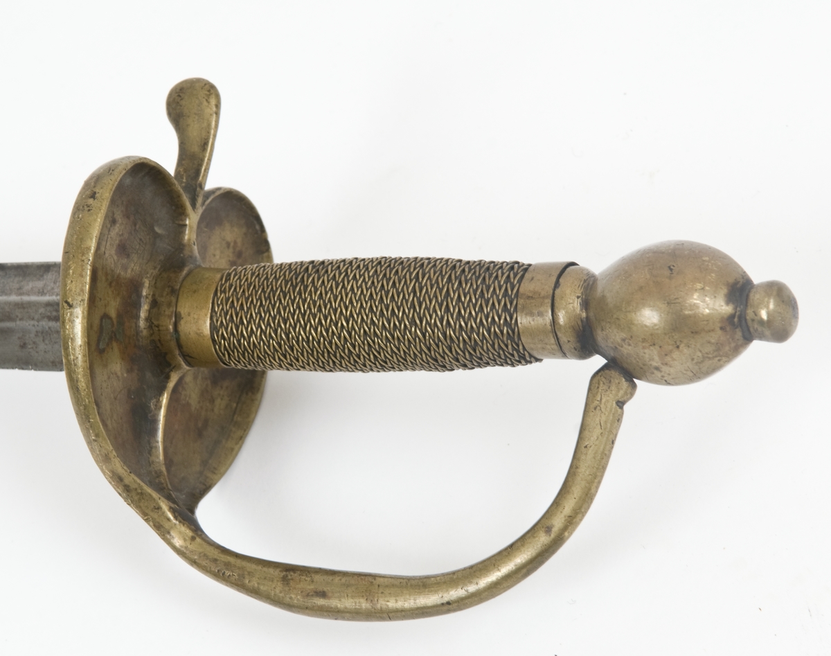 Offiserskorde dansk/norsk modell 1710. Messingfeste. Plommeformet knapp med nitteknapp. Hjerteformede parerplater, Fremre parerstang går over i håndbøylen som er tappet inn i knappen, rett bakre parerstang som ender i en flattrykt dråpe. Tvunnet messingtråd grep med messingbånd avslutning oppe og nede. Tveegget, sekskantet, rett klinge. Christian VI kronede speilmonogram på begeg sider av klingen.