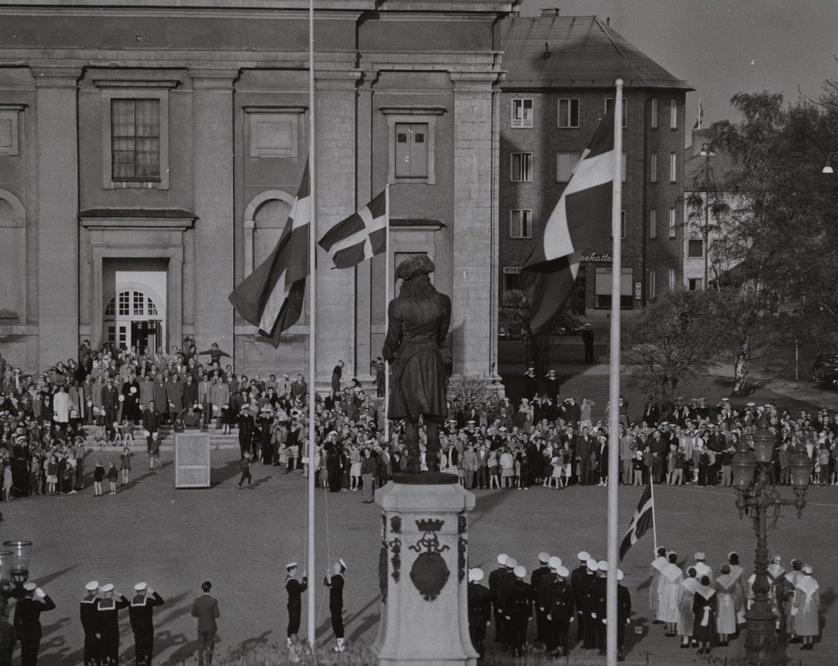 En folksamling har samlats utanför Fredrikskyrkan på Stortorget i Karlskrona och tittar på när två flottister hissar eller halar den svenska flaggan. Vid Karl XI:s staty står uniformsklädda militärer uppställda, varav några gör honör. I bakgrunden bakom kyrkan skymtar militärhemmet.