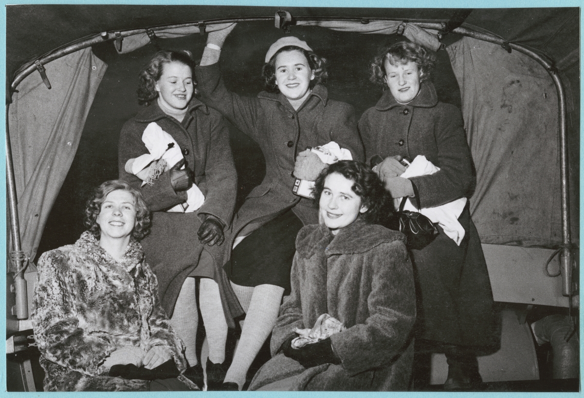 Fem unga kvinnor står och sitter i vad som ser ut som ett bilflak. Det är årets Lucia med tärnor, men på bilden har de fortfarande på sig sina vardagskläder och kappor, Luciakläderna håller de i famnen.