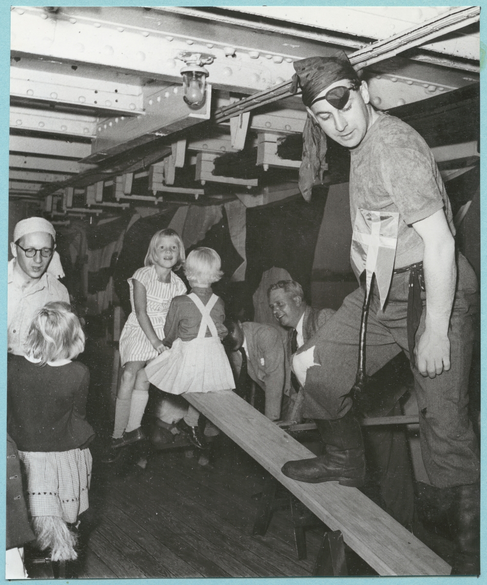 Vid barnbjudningen ombord på H.M.S. Montclare roades och trakterades barnen av till pirater utklädda besättningsmän. En besättningsman i piratkläder står på mitten av en gungbräda medan två flickor sitter på den ena änden. I bakgrunden syns ytterligare män och barn. Från engelska besöket i Karlskrona den 9-14 september 1951.