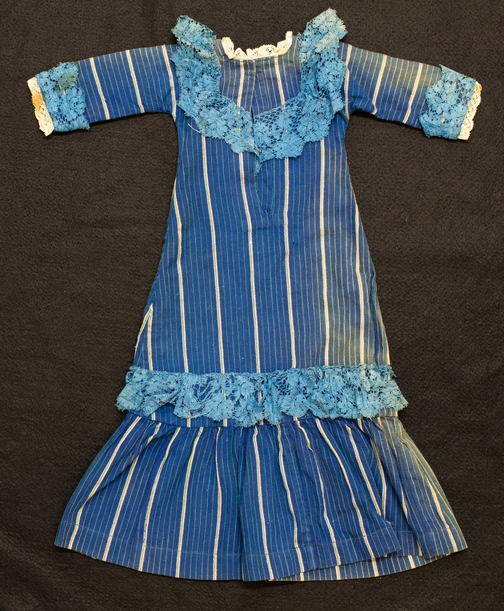 Blå bomullsklänning med vita ränder, blå spets runt halsringning och kjol, blekt fram. L. 52 cm.
Ingår i en samling dockkläder

Se VM 08 676.
