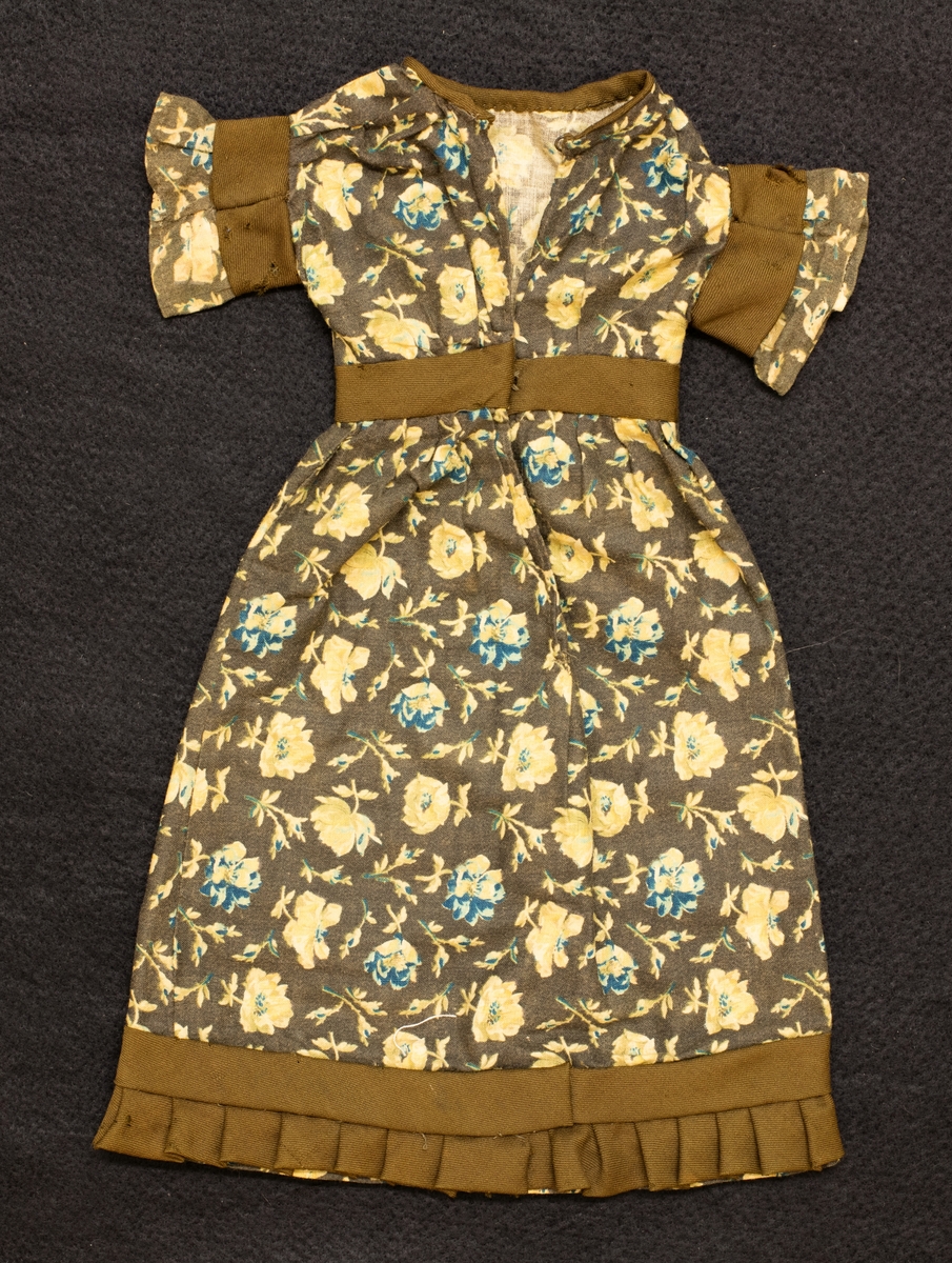Brun bomullsklänning med gula-blå blommor tryckta på tyget. Bruna band, kypert, i midjan, på ärmarna och som en volang längst ner på kjolen. L. 33 cm.