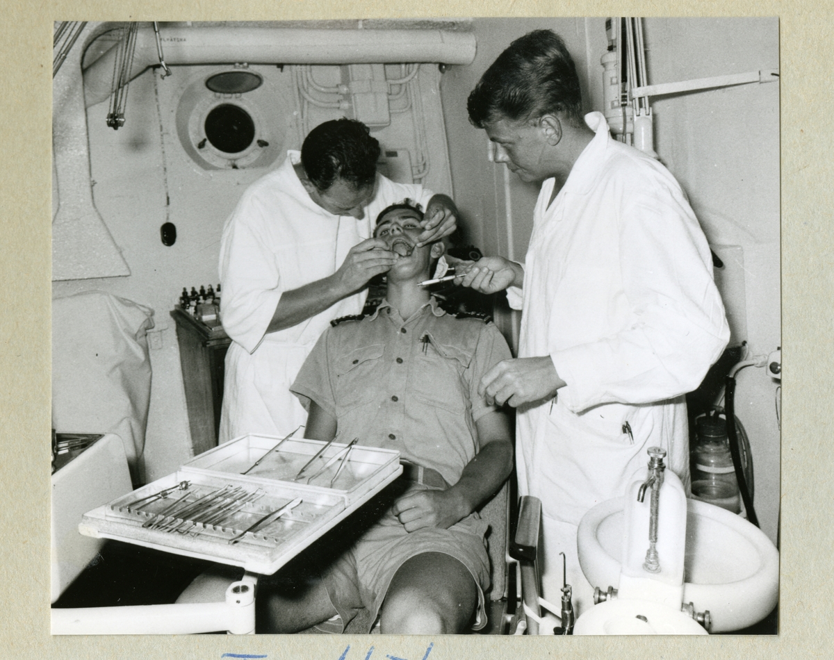 Bilden föreställer en besättningsmedlem som blir undersökt av tandläkare i vita rockar ombord på minfartyget Älvsnabben. Bilden är tagen under långresan 1966-1967.