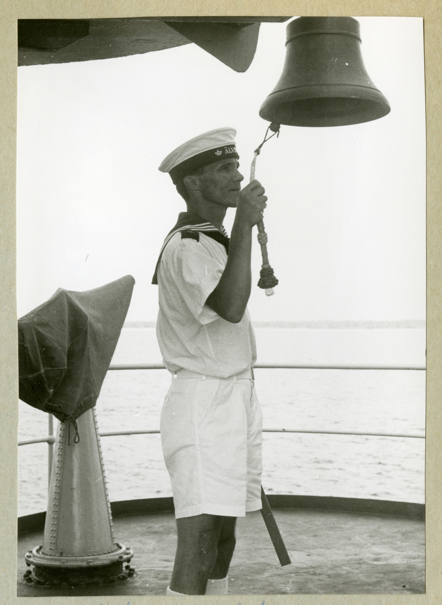 Bilden föreställer en besättningsman ombord på minfartyget Älvsnabben som slår i skeppsklockan iförd sommaruniform under långresan 1966-1967.
