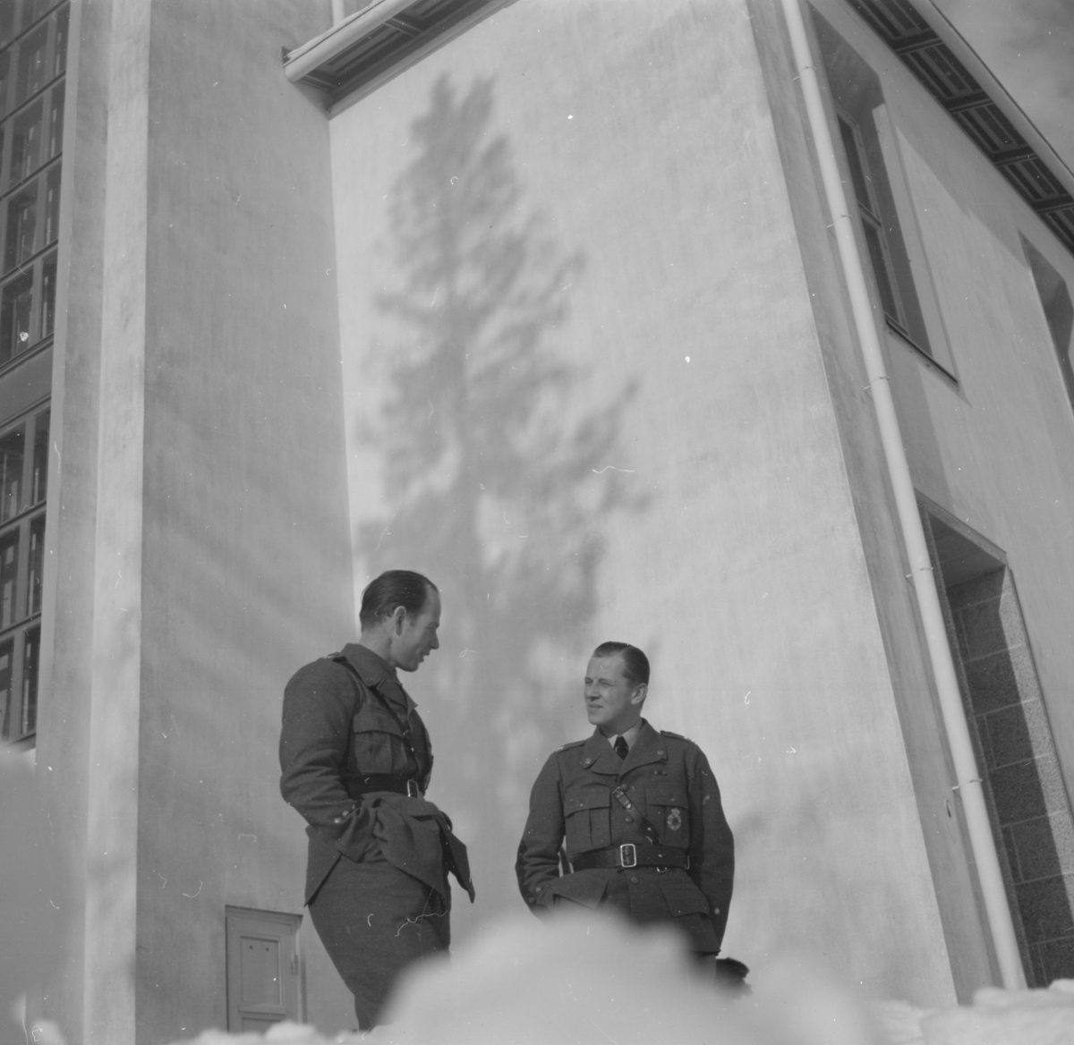 Två militärer tillhörande Svenska frivilligflottiljen i Finland, F 19, samtalar vid en byggnad. Snödriva i förgrunden.