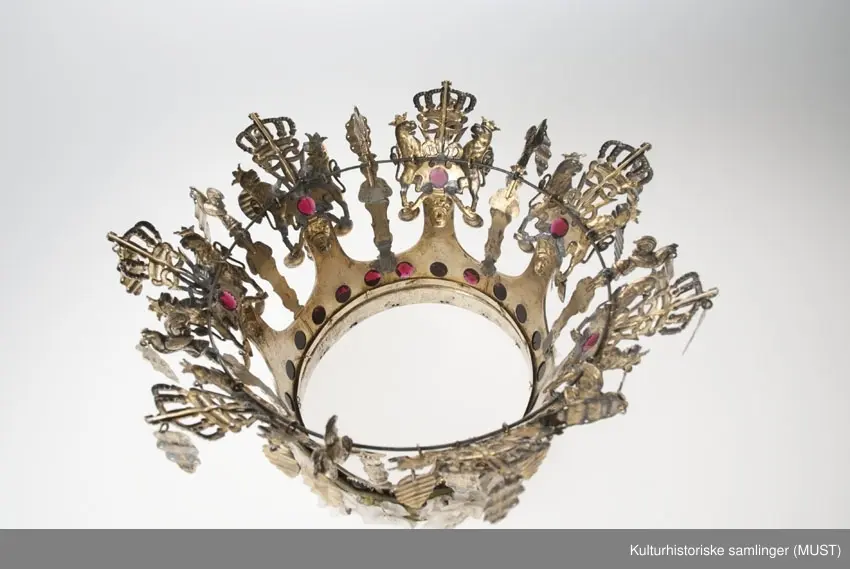 Brudekrone av sølv, forgylt. Nederst en glatt, innvendig stoppet ring, over
kronens 7 takker som munner ut i  Christian VII kronede monogram holdt av 2 løver,
og hver prydet med 4 røde glasstene og 1 mannshode med turbuan som holder et riflet
"løv". Til monogrammene er dessuten festet 3 "løv" og 2 skåler. Mellom
takkene en fløytespillende herme og en hane holdende et "løv". All
dekorasjon er presset og hvert takkestykke er arbeidet for seg og hengslet
til de øvrige. Det hele er avstivet med en messingtråd trukket gjennom
fastloddede øier på baksiden. Stemplet LH = Lauritz Hammer.1862.