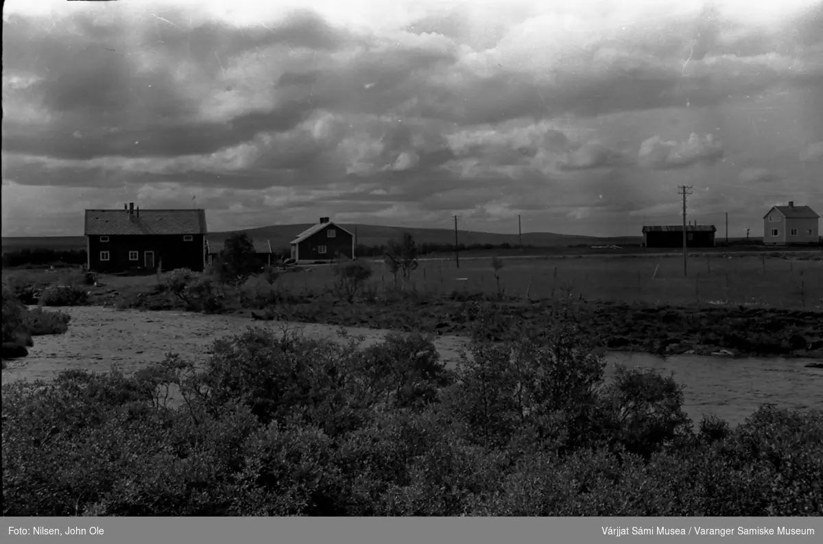 To bolighus med tilhørende bygninger, elv i forgrunnen. Ukjent sted, muligens i nærheten av Kautokeino. Juli 1967.