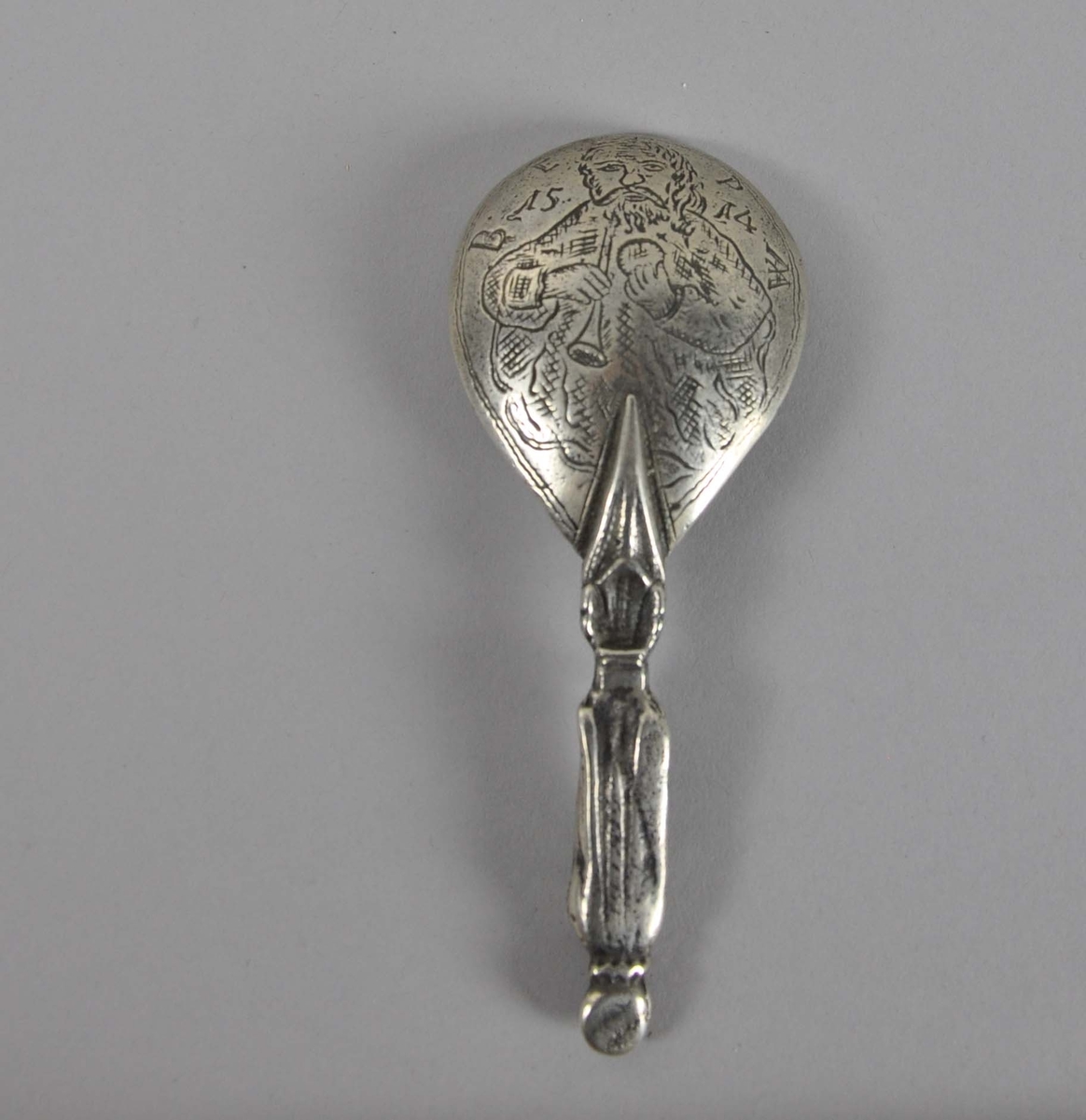 Rikt dekorert skje, av sølv. Skaftet har ikonisk motiv av mann med stav og bok. På baksiden av skjehodet er det inngravert innskrift rundt en mann med trompet og ball.