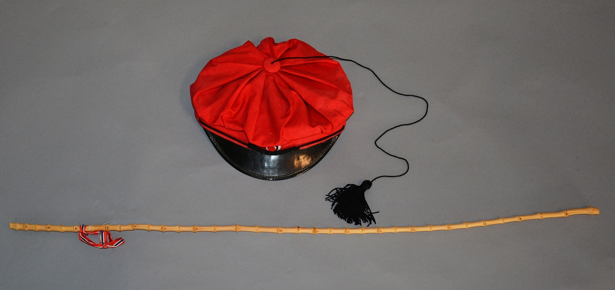 Rød russelue med smal skygge foran, tøyet samlet i legg som går sammen i en spiss på toppen, her er festet en knapp hvorpå det er festet en tråd som ender i dusk. Foret.