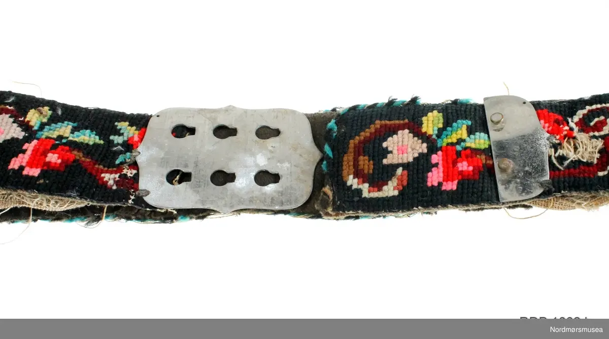Mannsbelte brodert på stramei med blomster- og bladmotiv i mange farger. Beltet er fôret med svart ullstoff og er påsydd ei sølvfarga metallspenne. Innleggsfor i lin. Beltet er svært slitt og fôret er revnet.