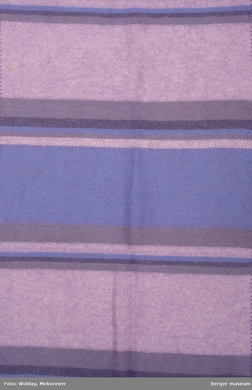 En pleddprøve i stripemønster. Prøven har jarekanter på kortsidene, de to langsidene har sydde kanter. Mønsteret karakterisereres av tverrgående felt og striper i forskjellge farger. Fargeholdningen er duse lilla toner.