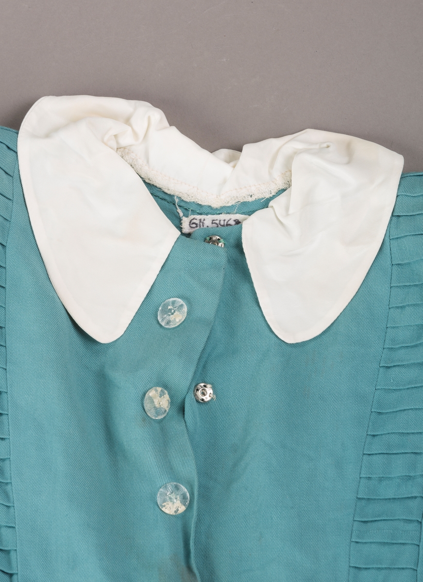 Form: Innsvingt liv, korte ermer, plissert skjørt, fastsydd belte.
Krave.
Bærestykket har vertikale stolper på hver side fra skulder ned til liv, med sydde legg.
Åpning foran med trykknapper. 3 pynteknapper utenpå.