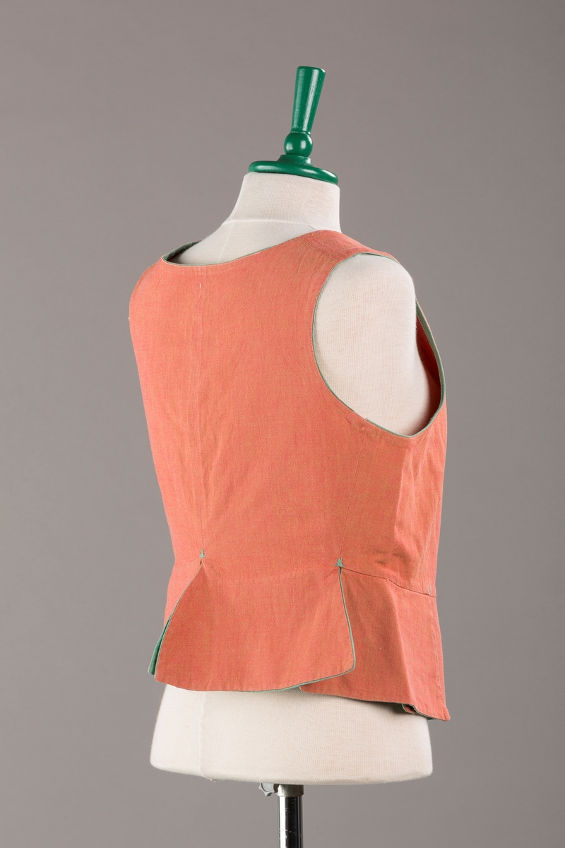 Rustrødt liv med grønn kant og fór, seks skjøter. Håndsydd forsterkning i kontrastfarge øverst i skjøtesplittene. Lukkes med tinnhekte og silkesnor.