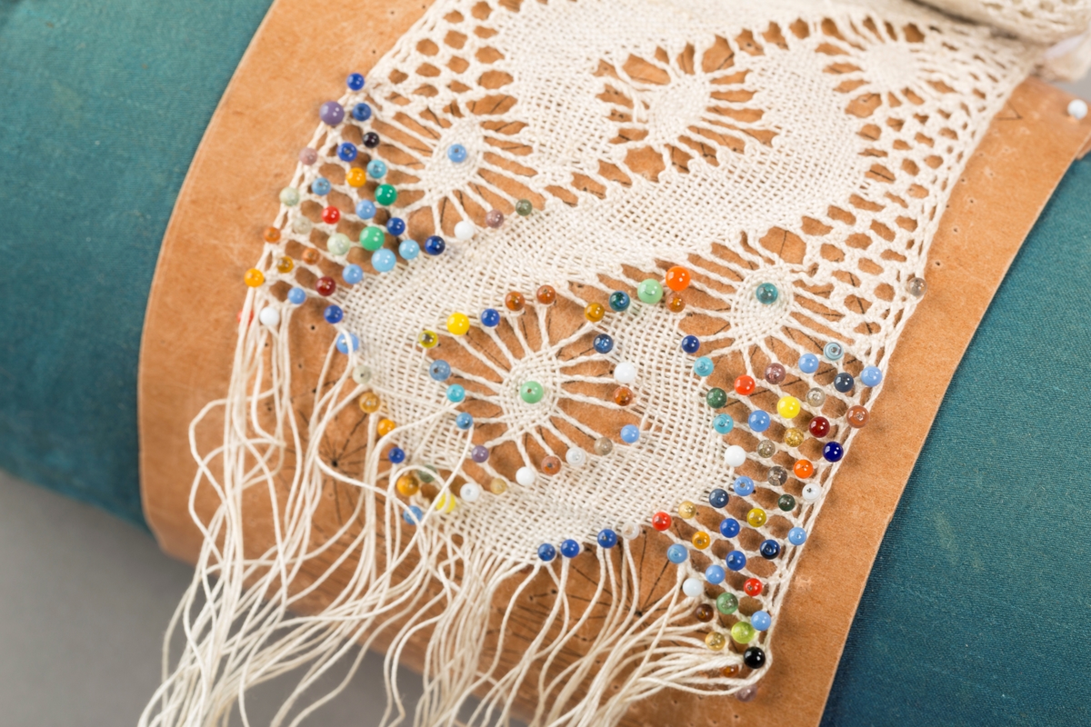 Form: Sylinderformet "pute" hvor stivt pappstykke rundt puta viser mønsteret. 
Kniplingsstykke under arbeid påspent med knappenåler
Pinner medkniplingstråd