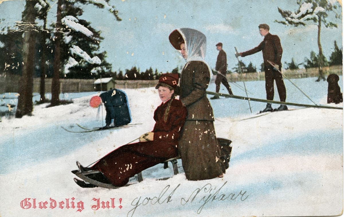 Julekort. Jule- og nyttårshilsen. Vintermotiv med vinteraktiviteter på snødekt jorde. To kvinner med kjelke og to menn samt barn på ski. Stemplet 22.12.1908.