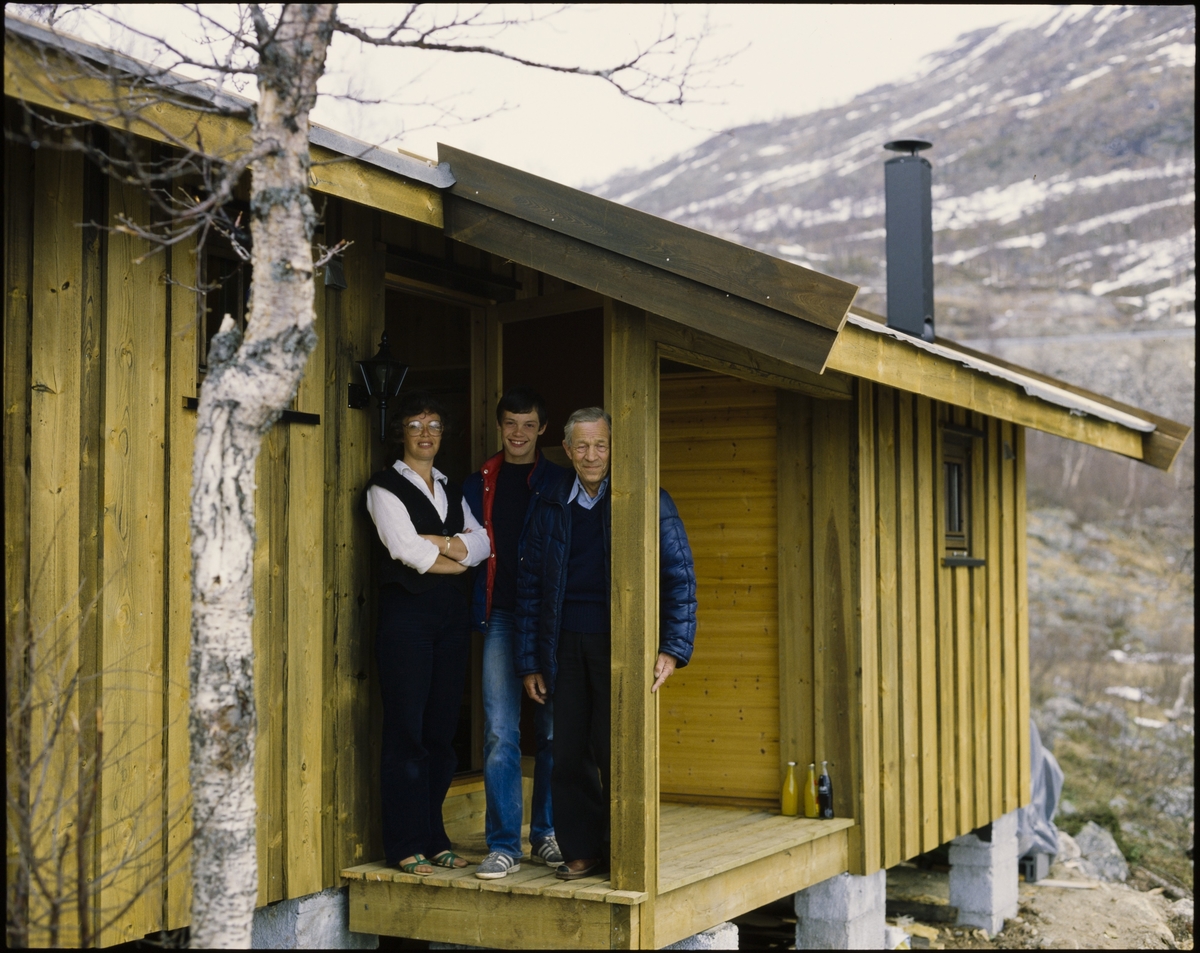 Tre personer smiler til fotografen ved inngangspartiet til en av Norsk Hydro sine hytter på Vågslid.