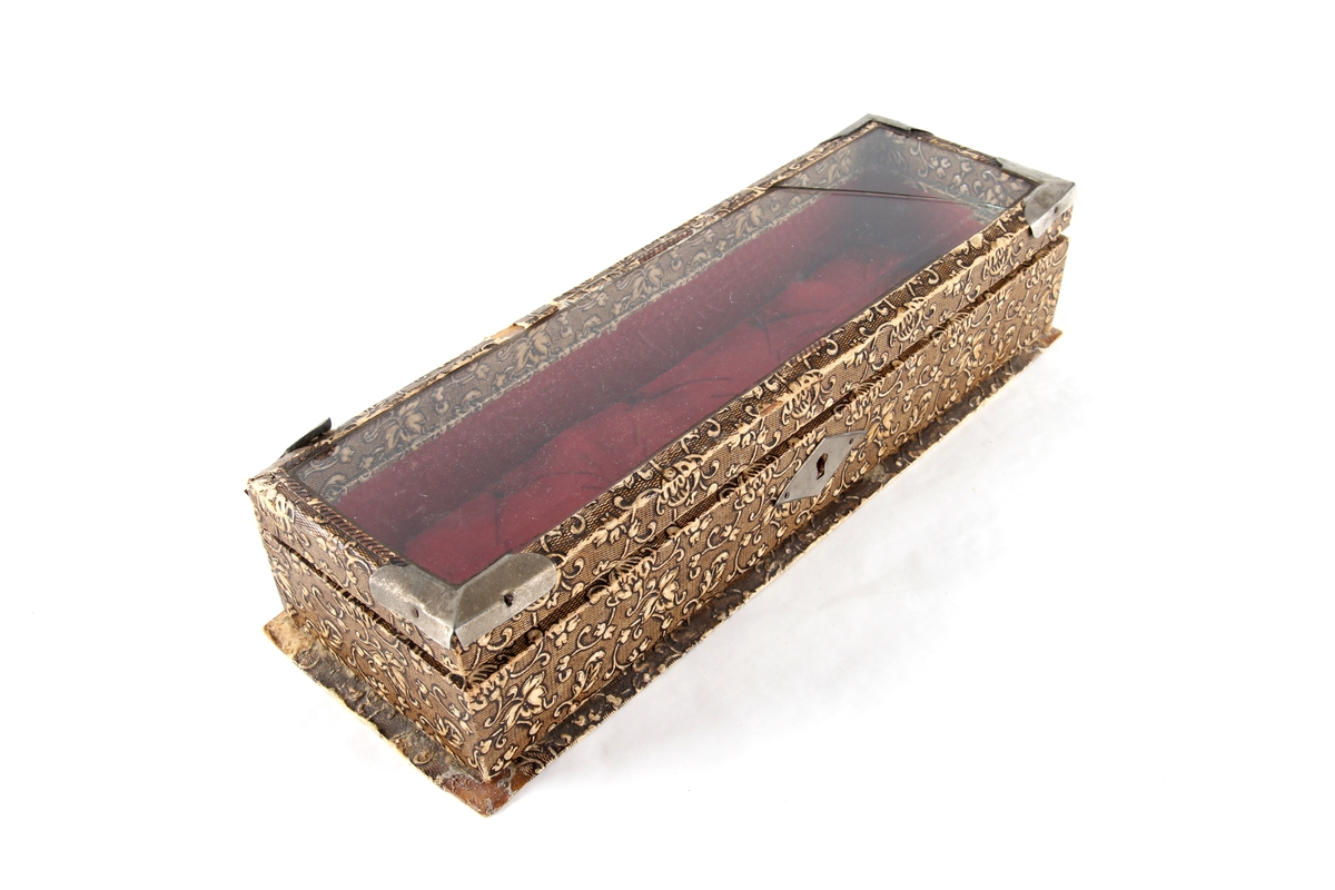 Avlangt smykkeskrin med hengsler og lokk. Lås og hjørnebeslag i metall. Lokket har et glassvindu. Foret med tekstil. Brukt til oppbevaring av små smykkegjenstander og en liten nøkkel.