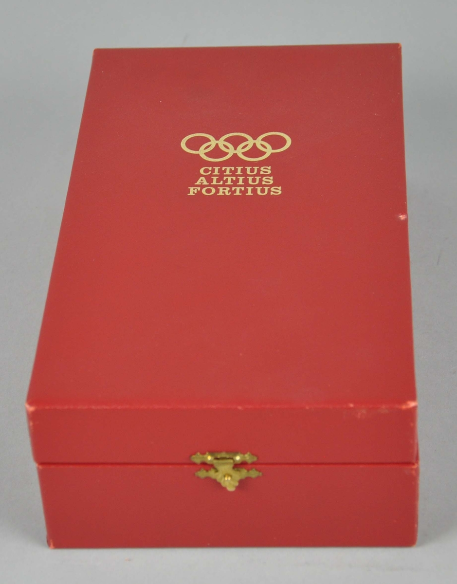 Olympisk torso på sokkel, med de olylmpiske ringer øverst. Torsoen ligger i et rødt etui, med et hefte som beskriver gjenstanden.