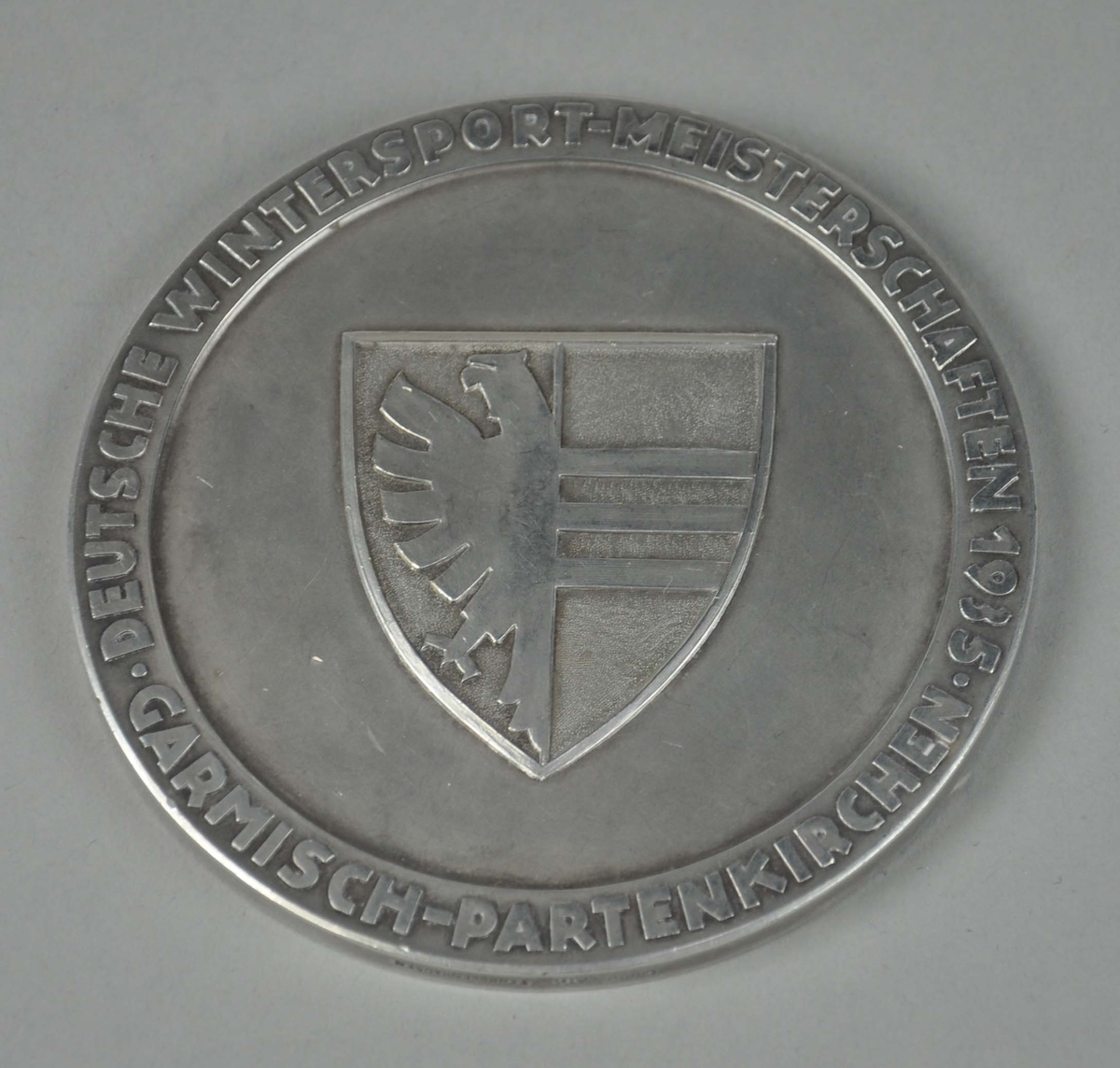 Rund sølvmedalje med byvåpenet for Garmisch-Partenkirchen i midten og tekst rundt kanten. På baksiden hakekors med tekst rundt kanten.